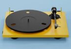 Platine vinyle Pro-Ject E1.2 BT en finition Satin Golden Yellow vue de trois quarts, plateau aluminium noir 730 g, bras aluminium 8,6 pouces avec cellule Pick it MM E et charnières dorées sur fond bleu clair