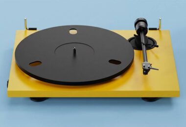 Platine vinyle Pro-Ject E1.2 BT en finition Satin Golden Yellow vue de trois quarts, plateau aluminium noir 730 g, bras aluminium 8,6 pouces avec cellule Pick it MM E et charnières dorées sur fond bleu clair