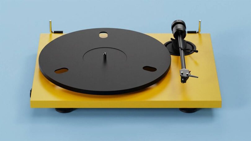 Platine vinyle Pro-Ject E1.2 BT en finition Satin Golden Yellow vue de trois quarts, plateau aluminium noir 730 g, bras aluminium 8,6 pouces avec cellule Pick it MM E et charnières dorées sur fond bleu clair