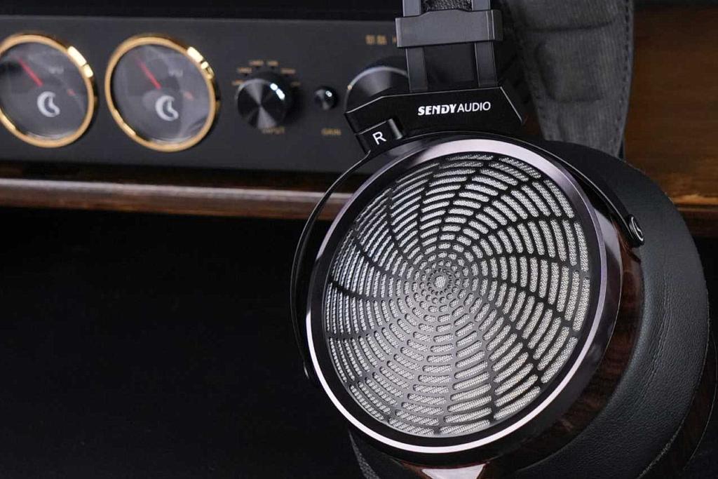 Gros plan sur la grille métallique à motif sunburst du casque planar Sendy Audio Apollo PRO et sa coque zébrano, devant un amplificateur casque à vumètres avec molettes de sortie et de gain