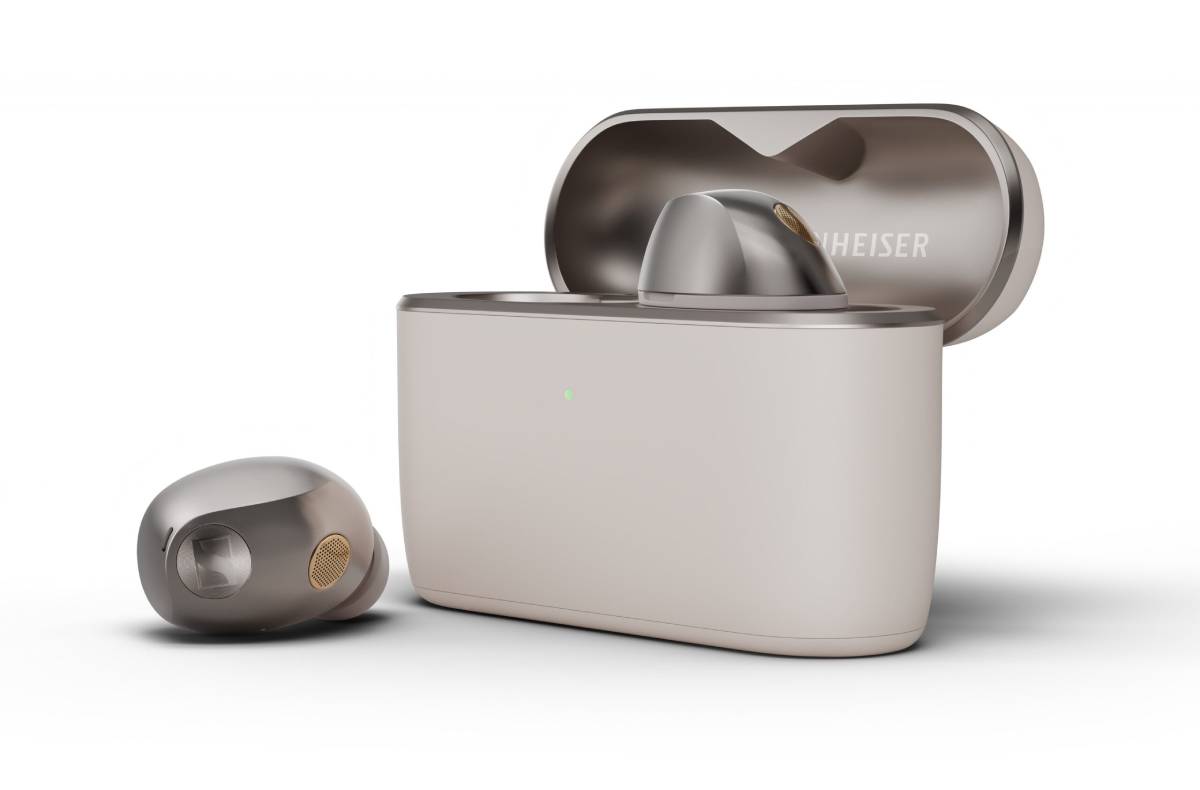 Boîtier de charge Sennheiser Momentum True Wireless 5 en finition crème mate, couvercle ouvert avec un écouteur métallisé logé dans son berceau, second écouteur posé devant montrant la plaque logo Sennheiser et la grille de microphone dorée, témoin de charge vert allumé