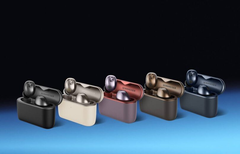 Écouteurs Sennheiser Momentum True Wireless 5 en finition Graphite posés devant leur boîtier de charge Qi au format vertical, avec le transducteur TrueResponse 7 mm et les grilles de microphones visibles sur la face externe des écouteurs