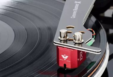 Cellule MC Vertere Ruby One à corps aluminium rouge anodisé montée sous un bras Vertere Groove Runner X, vis de fixation nickelées apparentes, câblage de headshell coloré et pointe diamant en contact avec le sillon d'un disque vinyle noir