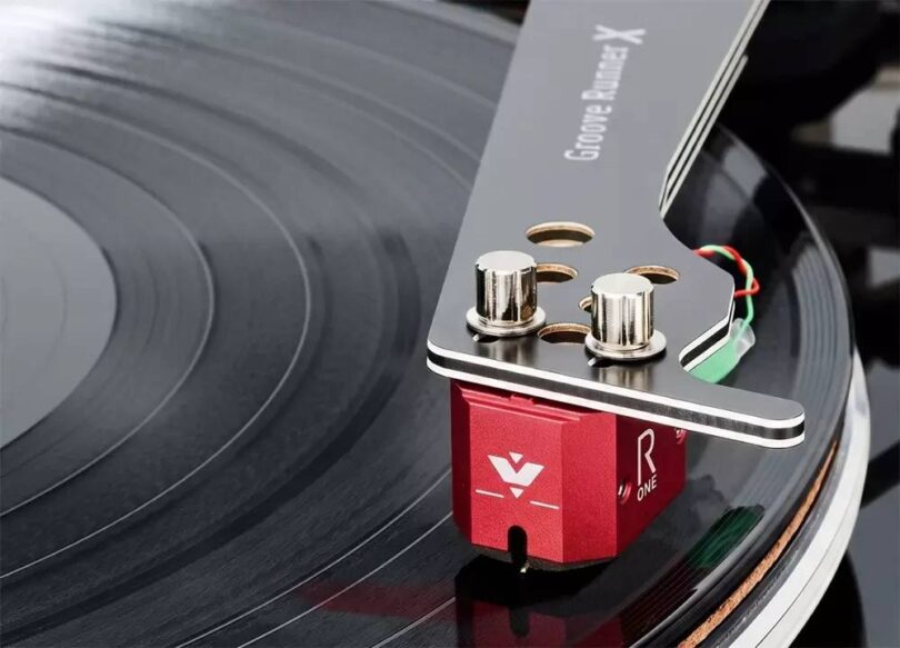 Cellule MC Vertere Ruby One à corps aluminium rouge anodisé montée sous un bras Vertere Groove Runner X, vis de fixation nickelées apparentes, câblage de headshell coloré et pointe diamant en contact avec le sillon d'un disque vinyle noir