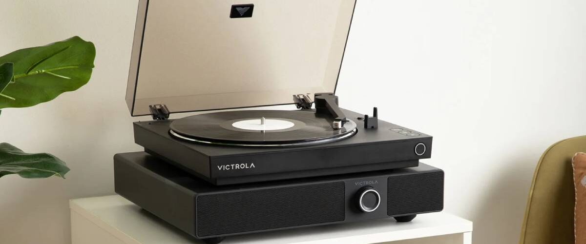 Enceinte soundbase Victrola Soundstage en finition noire posée sur un meuble blanc, avec une platine vinyle Victrola noire installée sur son plateau, capot fumé ouvert, bouton rotatif multifonction et grille frontale visibles dans un salon lumineux avec plante verte