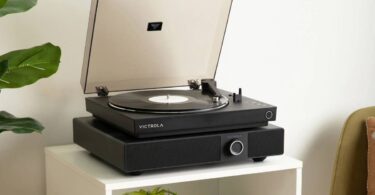 Enceinte soundbase Victrola Soundstage en finition noire posée sur un meuble blanc, avec une platine vinyle Victrola noire installée sur son plateau, capot fumé ouvert, bouton rotatif multifonction et grille frontale visibles dans un salon lumineux avec plante verte