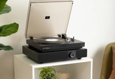 Enceinte soundbase Victrola Soundstage en finition noire posée sur un meuble blanc, avec une platine vinyle Victrola noire installée sur son plateau, capot fumé ouvert, bouton rotatif multifonction et grille frontale visibles dans un salon lumineux avec plante verte