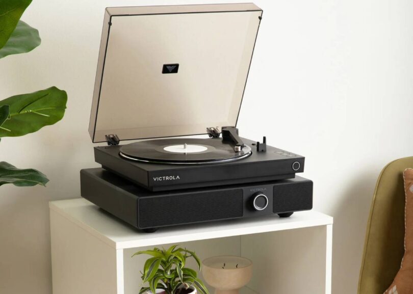 Enceinte soundbase Victrola Soundstage en finition noire posée sur un meuble blanc, avec une platine vinyle Victrola noire installée sur son plateau, capot fumé ouvert, bouton rotatif multifonction et grille frontale visibles dans un salon lumineux avec plante verte