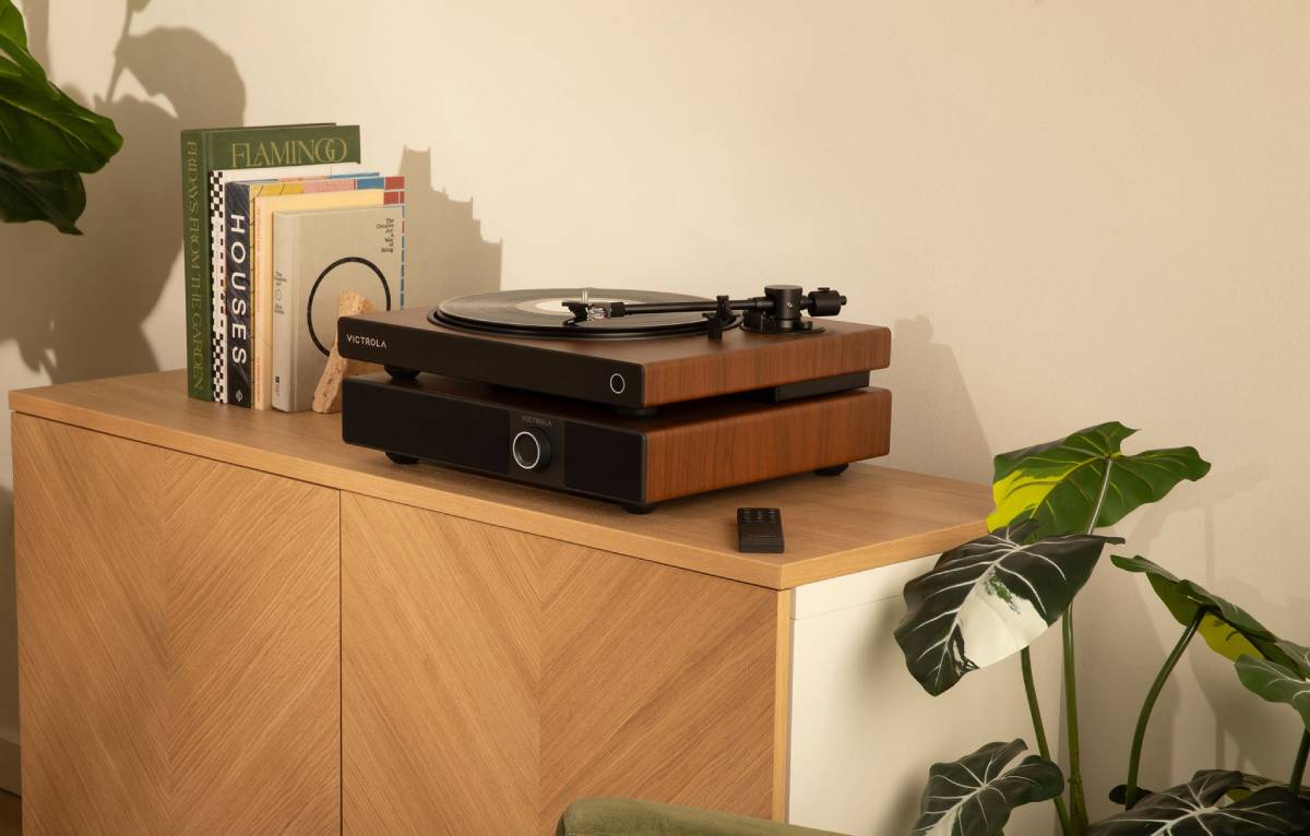 Victrola Soundstage en finition noyer surmontée d'une platine vinyle Victrola assortie sur un buffet en chêne clair, télécommande infrarouge posée à côté, livres et plantes vertes en arrière-plan dans un intérieur contemporain
