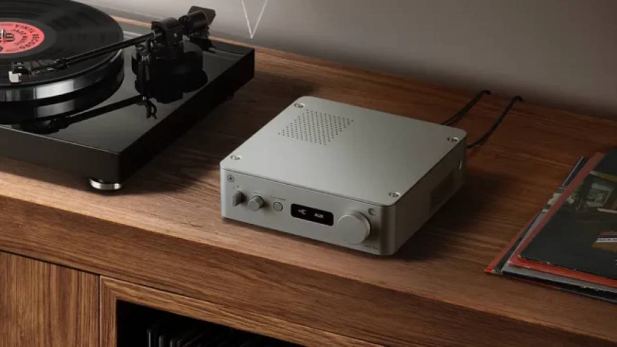 Amplificateur réseau Yamaha WXA-70A en finition argent posé sur un meuble en chêne massif à côté d'une platine vinyle noire en lecture, grille de ventilation ajourée sur le capot, écran affichant la source AUX et molette de volume, pochettes de disques vinyle à droite
