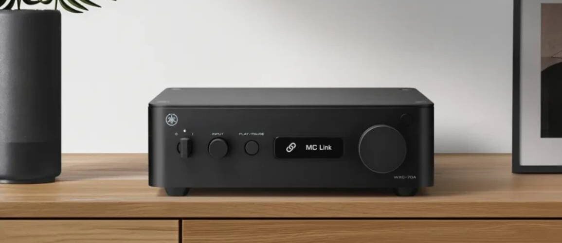 Yamaha WXC-70A en finition noire posé sur un meuble en bois clair, face avant avec interrupteur d'alimentation, sélecteur de source INPUT, touche PLAY/PAUSE, écran affichant MC Link et large molette de volume à droite