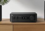 Yamaha WXC-70A en finition noire posé sur un meuble en bois clair, face avant avec interrupteur d'alimentation, sélecteur de source INPUT, touche PLAY/PAUSE, écran affichant MC Link et large molette de volume à droite