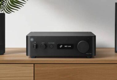 Yamaha WXC-70A en finition noire posé sur un meuble en bois clair, face avant avec interrupteur d'alimentation, sélecteur de source INPUT, touche PLAY/PAUSE, écran affichant MC Link et large molette de volume à droite