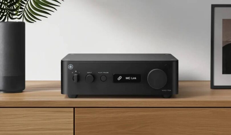 Yamaha WXC-70A en finition noire posé sur un meuble en bois clair, face avant avec interrupteur d'alimentation, sélecteur de source INPUT, touche PLAY/PAUSE, écran affichant MC Link et large molette de volume à droite