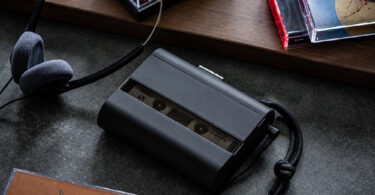 Baladeur cassette Amadana CP10 noir avec panneau avant fumé semi-transparent laissant voir la cassette, dragonne tressée noire, casque filaire rétro à coussinets en mousse grise et boîtiers de cassettes audio