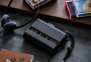 Baladeur cassette Amadana CP10 noir avec panneau avant fumé semi-transparent laissant voir la cassette, dragonne tressée noire, casque filaire rétro à coussinets en mousse grise et boîtiers de cassettes audio