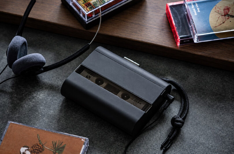 Baladeur cassette Amadana CP10 noir avec panneau avant fumé semi-transparent laissant voir la cassette, dragonne tressée noire, casque filaire rétro à coussinets en mousse grise et boîtiers de cassettes audio