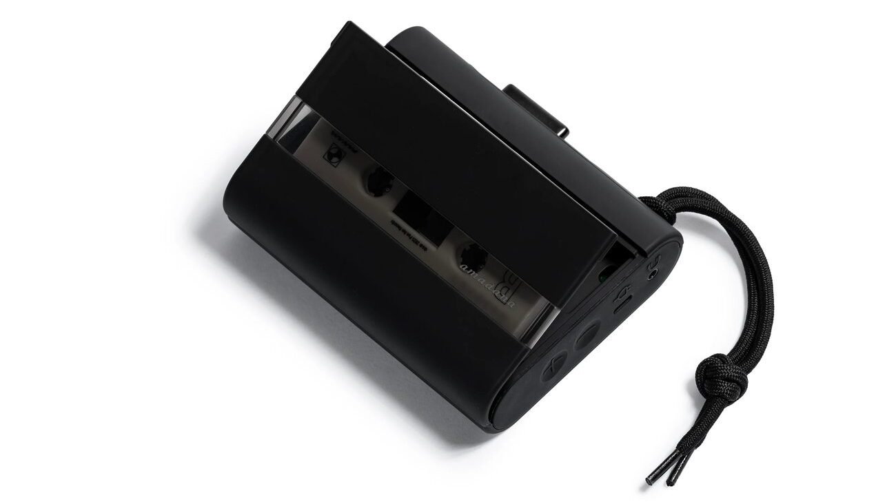 Baladeur cassette Amadana CP10 noir mat avec sa trappe entrouverte, cassette chargée visible derrière le panneau fumé semi-transparent, boutons de volume, port USB-C et prise jack sur la tranche, dragonne tressée noire
