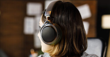 Femme de profil portant le casque ouvert sans fil Audio-Technica ATH-AL5NC, avec grille hexagonale métallique, arceau textile, logo Audio-Technica et accents bronze, dans un intérieur chaleureux