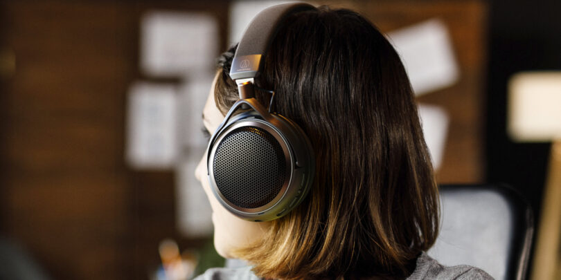 Femme de profil portant le casque ouvert sans fil Audio-Technica ATH-AL5NC, avec grille hexagonale métallique, arceau textile, logo Audio-Technica et accents bronze, dans un intérieur chaleureux