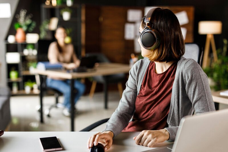 Femme portant le casque ouvert sans fil Audio-Technica ATH-AL5NC à son bureau, devant un ordinateur portable et un smartphone, se tournant vers une collègue dans un espace de travail partagé