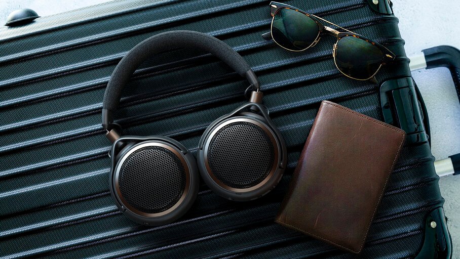 Casque ouvert sans fil Audio-Technica ATH-AL5NC à coques pivotées à plat, grilles hexagonales et accents bronze, posé sur une valise noire à côté de lunettes de soleil et d'un porte-passeport en cuir