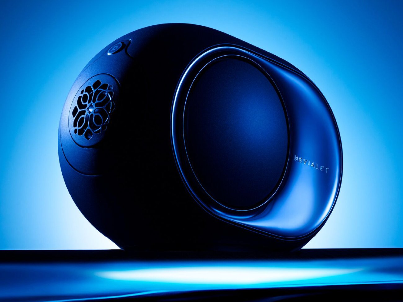 Gros plan de l'enceinte Devialet Phantom Ultimate 98 dB Pulse Blue en bleu RAL 5011, flasque latérale polie miroir gravée du logo Devialet, grille cosphérique ajourée et bouton tactile supérieur