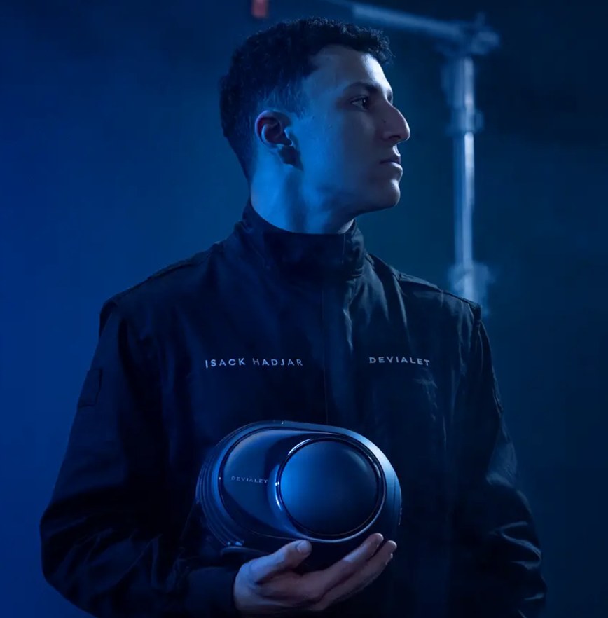 Le pilote de Formule 1 Isack Hadjar en veste noire brodée Devialet tenant une enceinte Devialet Phantom Ultimate 98 dB Pulse Blue à flasque miroir, dans un studio éclairé en bleu