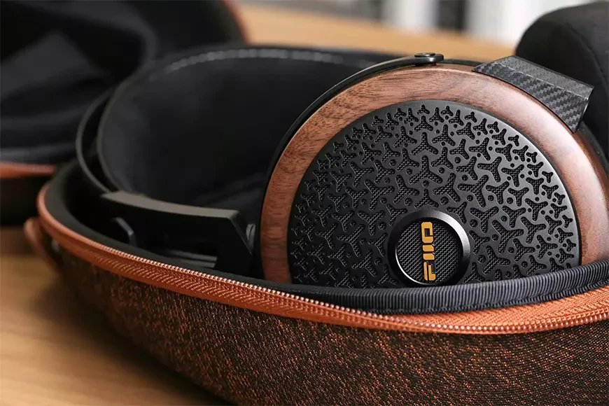 Gros plan sur la coque en noyer noir massif du casque planaire FiiO FT15, sa grille ajourée noire à motif organique, son logo doré et la pièce en fibre de carbone de l'arceau, rangé dans son étui rigide en tissu cuivré zippé