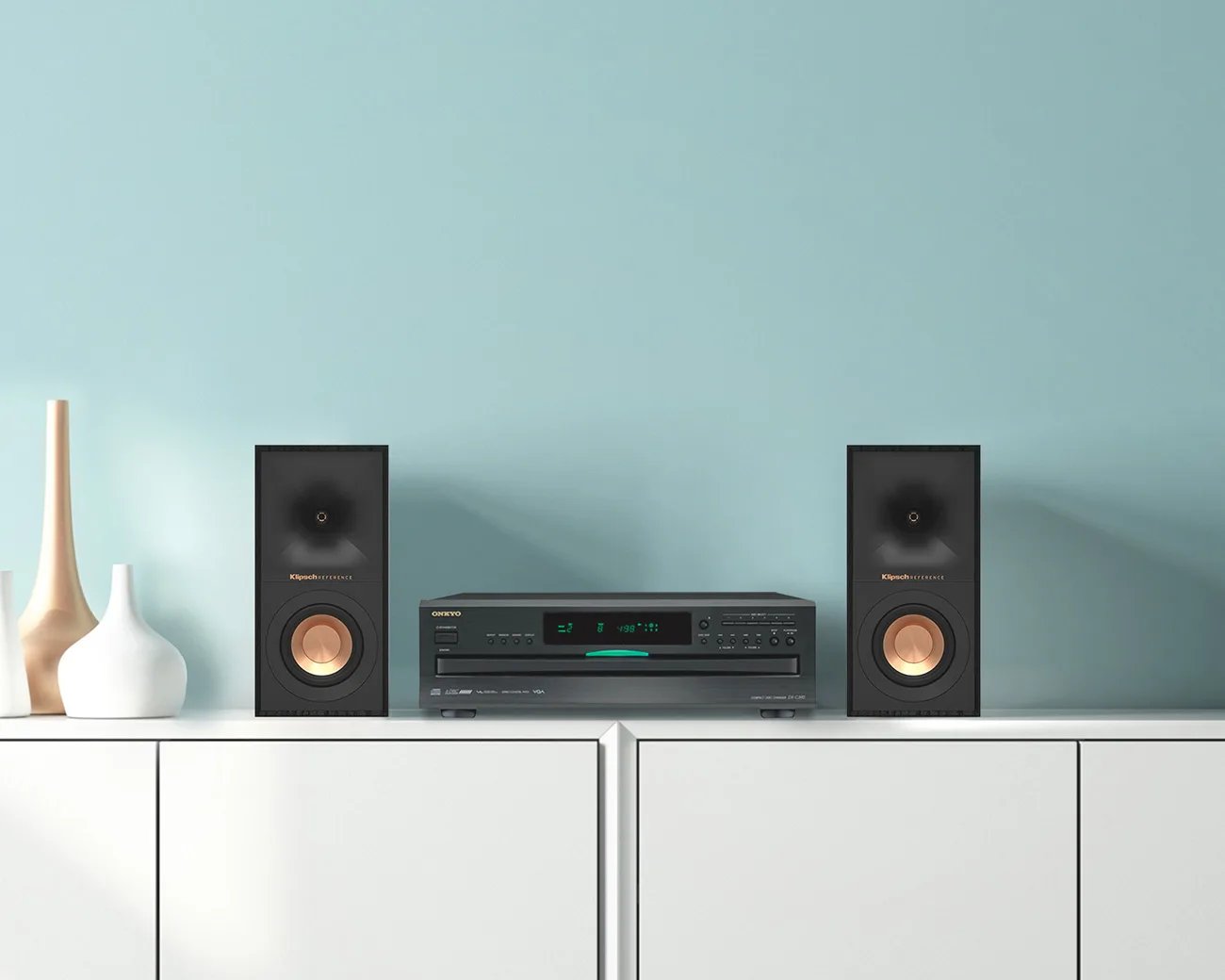 Changeur CD Onkyo DX-C390 noir, tiroir fermé et afficheur allumé, posé sur un meuble blanc entre deux enceintes bibliothèque Klipsch Reference, devant un mur bleu pastel
