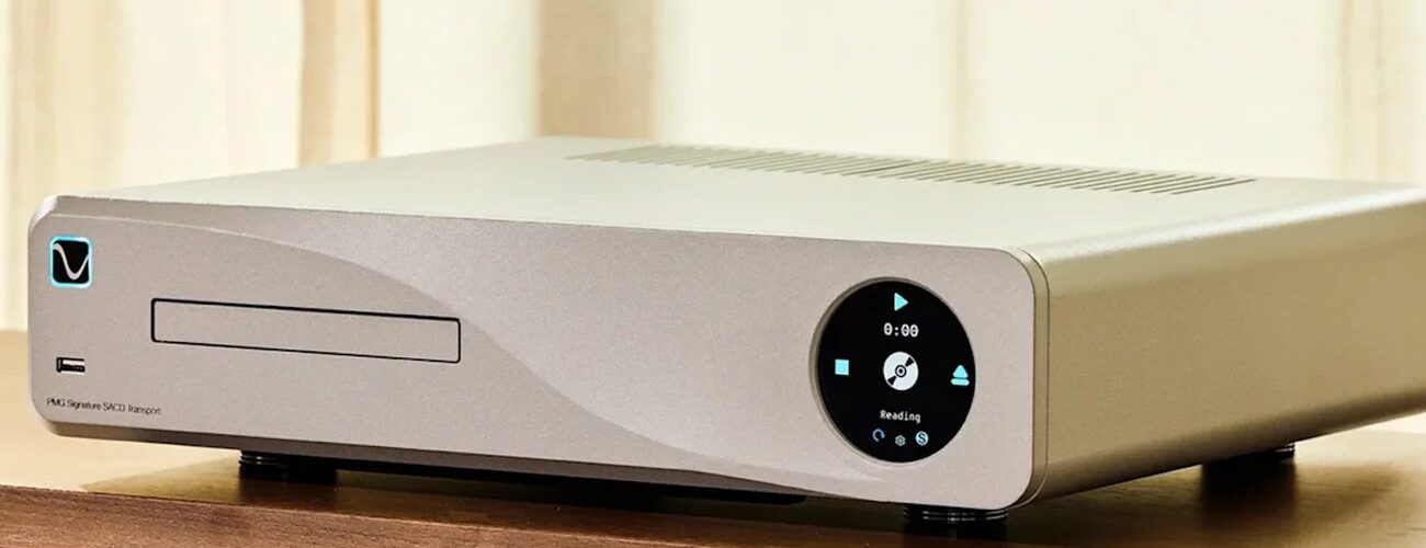 PS Audio PMG Signature SACD Transport en finition argent posé sur un meuble bois, tiroir de chargement CD et SACD à gauche, écran circulaire de commande à droite affichant Reading, pieds découplants cylindriques et châssis aluminium usiné