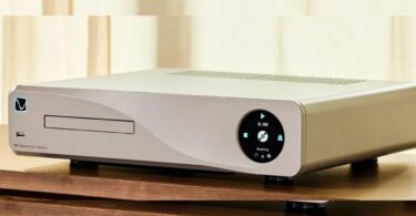 PS Audio PMG Signature SACD Transport en finition argent posé sur un meuble bois, tiroir de chargement CD et SACD à gauche, écran circulaire de commande à droite affichant Reading, pieds découplants cylindriques et châssis aluminium usiné