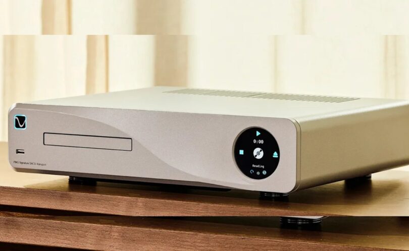 PS Audio PMG Signature SACD Transport en finition argent posé sur un meuble bois, tiroir de chargement CD et SACD à gauche, écran circulaire de commande à droite affichant Reading, pieds découplants cylindriques et châssis aluminium usiné