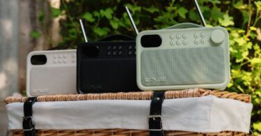 Trois radios portables Pure Bloom DAB+ et Bluetooth en coloris Sable, Noir et Sauge posées sur un panier de pique-nique en osier, avec antennes télescopiques, écran couleur, touches de présélection et façade en tissu