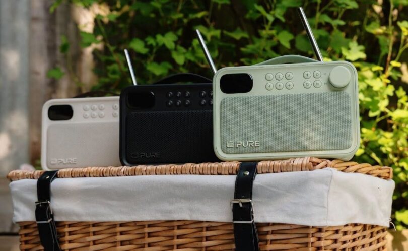 Trois radios portables Pure Bloom DAB+ et Bluetooth en coloris Sable, Noir et Sauge posées sur un panier de pique-nique en osier, avec antennes télescopiques, écran couleur, touches de présélection et façade en tissu