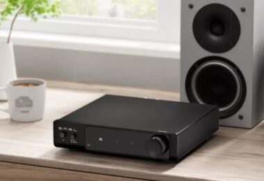 Amplificateur de puissance SMSL RAW-MHA1 noir posé sur un meuble en bois à côté d'enceintes de bibliothèque blanches, face avant avec bouton de volume, sélecteur d'entrée XLR RCA et afficheur indiquant le mode BTL