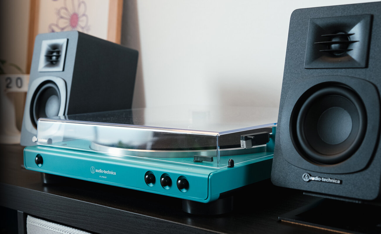 Façade bleu canard de la platine Audio-Technica AT-LP60XBT avec ses boutons Start, Stop et levée du bras, couvercle fermé, devant deux enceintes actives