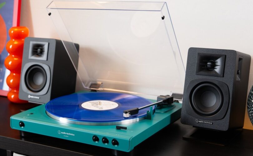 Platine vinyle Audio-Technica AT-LP60XBT en finition bleu canard avec couvercle ouvert et vinyle bleu sur le plateau, entourée de deux enceintes actives noires sur un meuble
