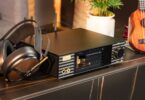 Cayin iD6 en finition noire posé sur un meuble, vu de trois quarts face : écran tactile, fenêtre sur les lampes 12AU7, molette de volume dorée, sorties casque en façade, antennes à l'arrière et casque ouvert posé à côté