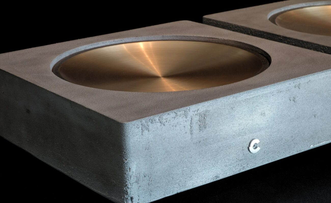 DAC Cinnamon Galle Model II : châssis en aluminium coulé au sable, dessus en bronze ciré à la main en forme de coupelle, logo Cinnamon en façade, second châssis en arrière-plan