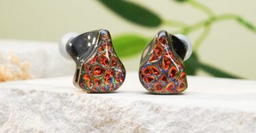 Paire d'intras DUNU PRS5 à coque en résine sombre et façades multicolores rouge et or, posée sur une pierre claire devant des feuilles de bambou