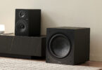 Caisson de basses Elipson Classic Sub 12 noir, haut-parleur de 30 cm en fibre de verre visible, posé au sol à côté d'un meuble TV noir et d'une enceinte bibliothèque dans un salon clair