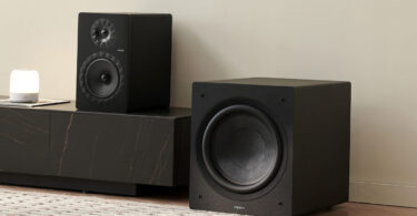 Caisson de basses Elipson Classic Sub 12 noir, haut-parleur de 30 cm en fibre de verre visible, posé au sol à côté d'un meuble TV noir et d'une enceinte bibliothèque dans un salon clair