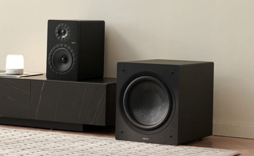 Caisson de basses Elipson Classic Sub 12 noir, haut-parleur de 30 cm en fibre de verre visible, posé au sol à côté d'un meuble TV noir et d'une enceinte bibliothèque dans un salon clair