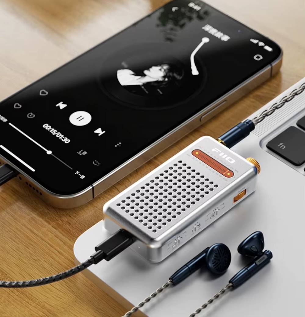 Radio FiiO RR13 argent utilisée en ampli casque : smartphone relié par un câble USB-C tressé au port USB-C de la radio, écouteurs branchés sur sa prise jack 3,5 mm, posée sur un ordinateur portable
