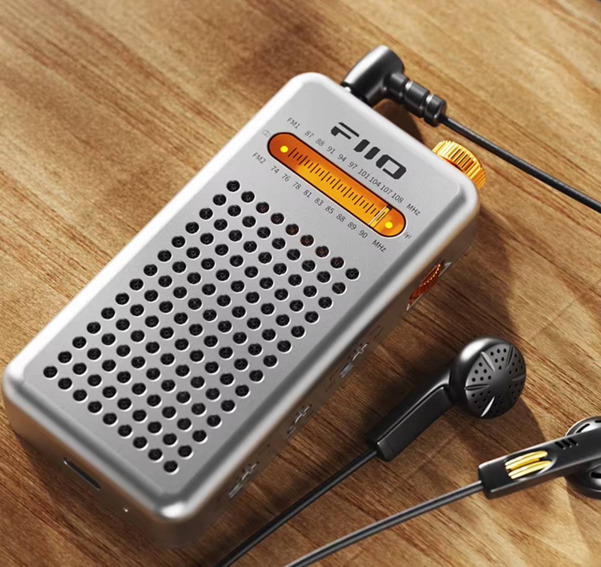 Radio FM de poche FiiO RR13 argent posée sur une table en bois, écouteurs noirs branchés par un jack 3,5 mm coudé, cadran orange rétroéclairé, molette d'accord dorée, grille de haut-parleur et port USB-C