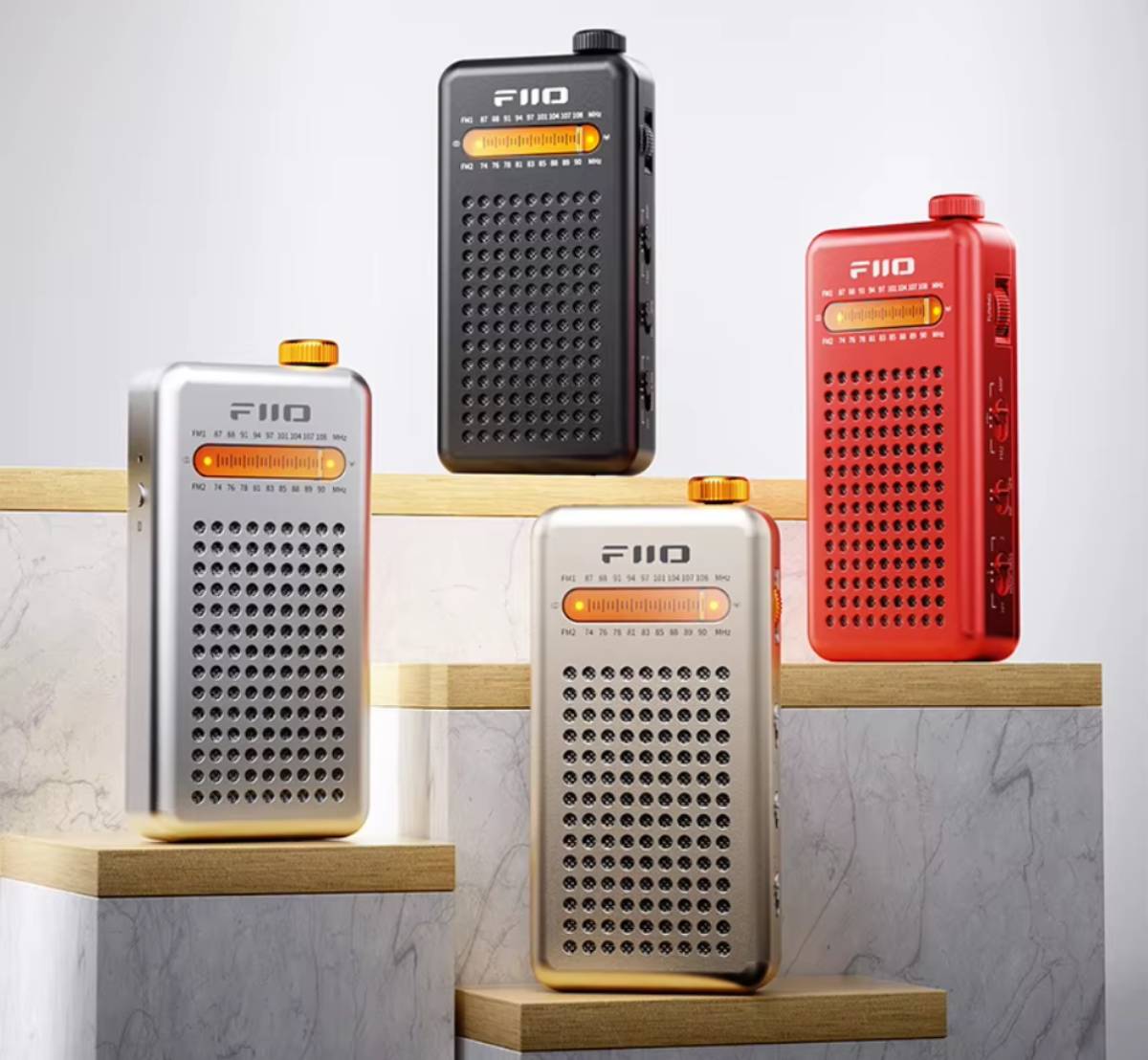 Les quatre coloris de la radio FM de poche FiiO RR13, noir, argent, rouge et or rétro, posés sur des socles en marbre et bois clair, avec cadran orange rétroéclairé, molette d'accord sur le dessus et grille de haut-parleur perforée