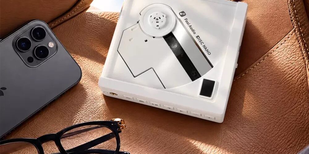 Lecteur CD portable Fosi Audio DISC Nano en finition blanche posé sur un siège en cuir fauve, disque à moitié apparent, rangée de boutons en façade, à côté d'un smartphone et de lunettes