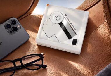 Lecteur CD portable Fosi Audio DISC Nano en finition blanche posé sur un siège en cuir fauve, disque à moitié apparent, rangée de boutons en façade, à côté d'un smartphone et de lunettes