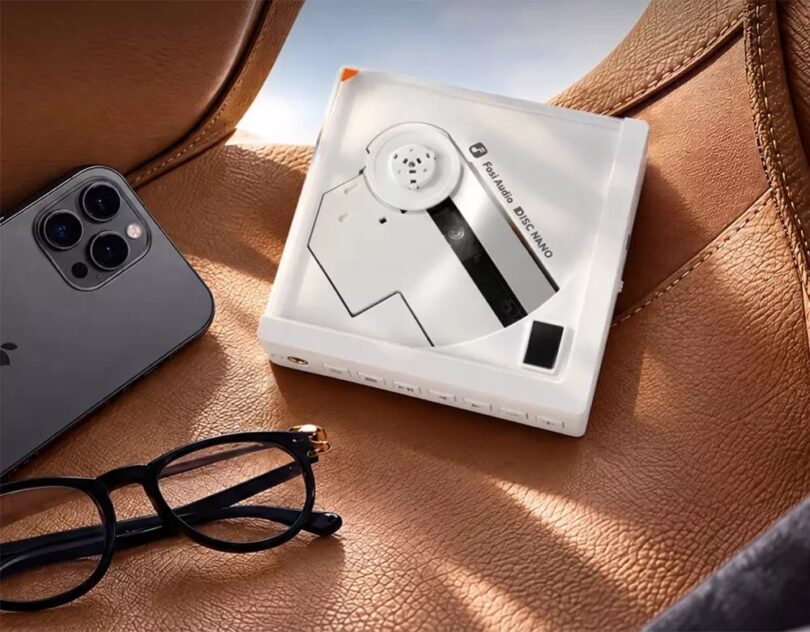 Lecteur CD portable Fosi Audio DISC Nano en finition blanche posé sur un siège en cuir fauve, disque à moitié apparent, rangée de boutons en façade, à côté d'un smartphone et de lunettes